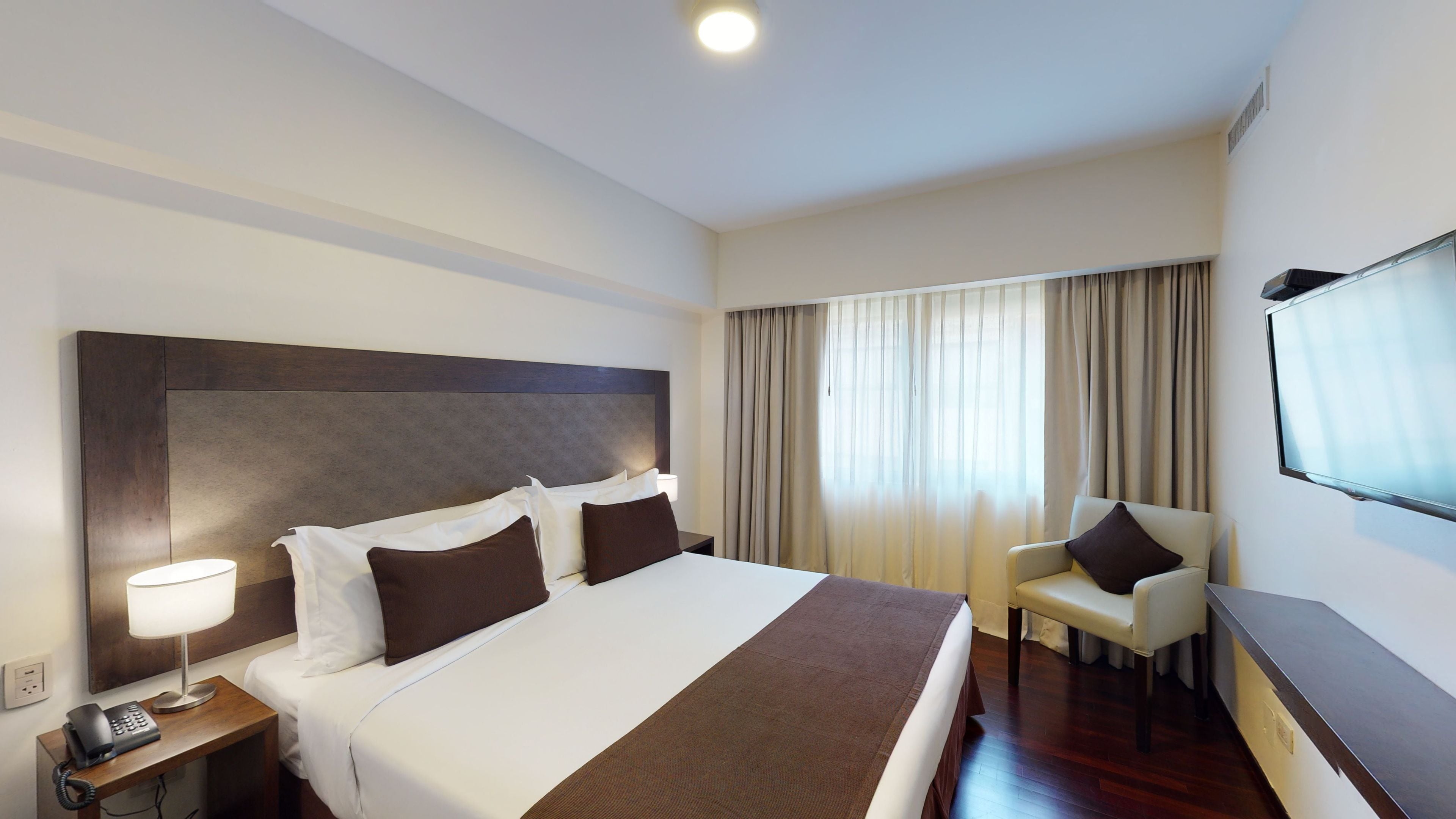 Habitación de hotel con una cama grande, un escritorio, un televisor de pantalla plana, una silla y una mesita de noche con lámpara y teléfono.