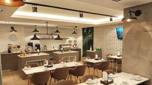 Un espace repas moderne avec plusieurs tables et chaises, un coin cuisine équipé d’appareils électroménagers et un mur en briques sur lequel est accrochée une télévision.