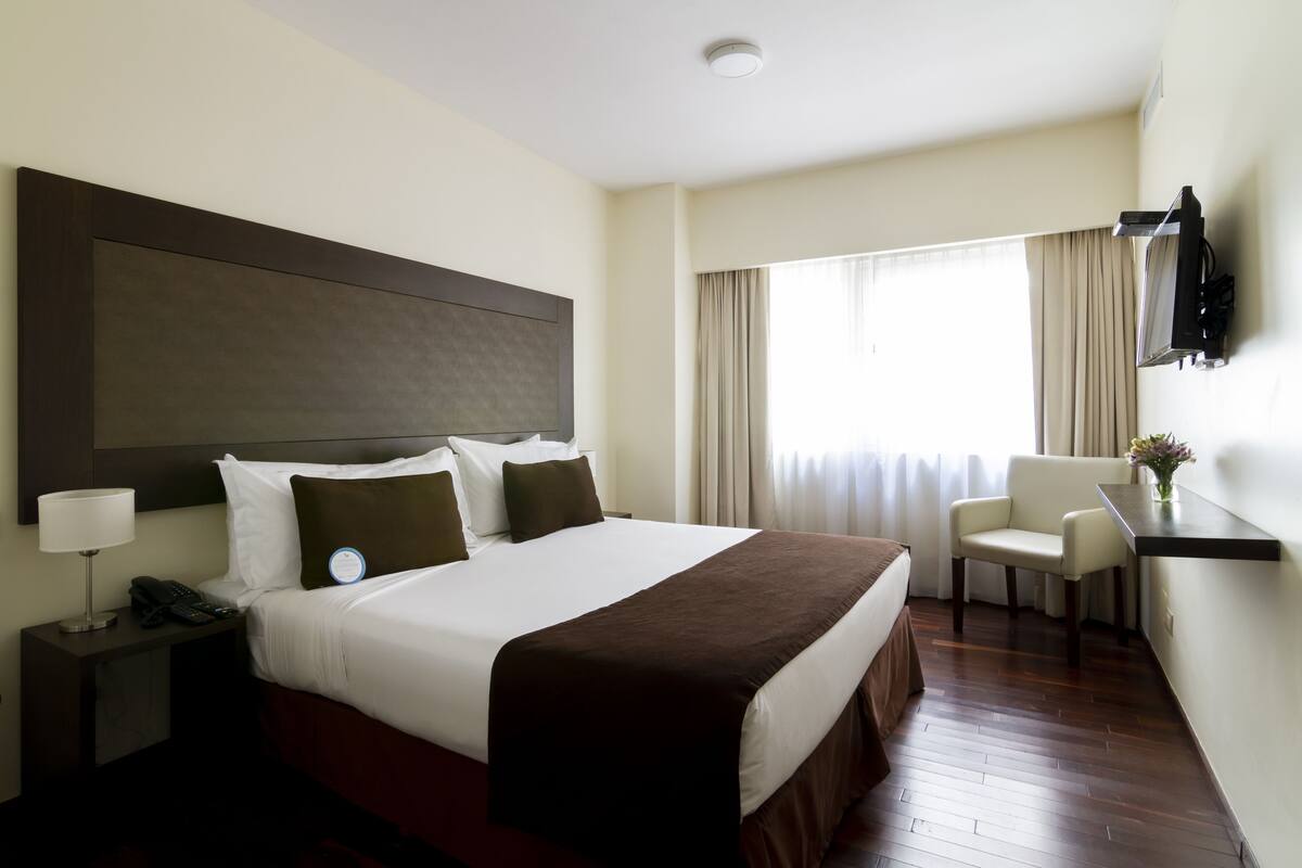 Ein Hotelzimmer mit einem großen Bett, einem Nachttisch mit Lampe, einem Sessel, einem an der Wand befestigten Fernseher und einem Fenster mit Vorhängen.