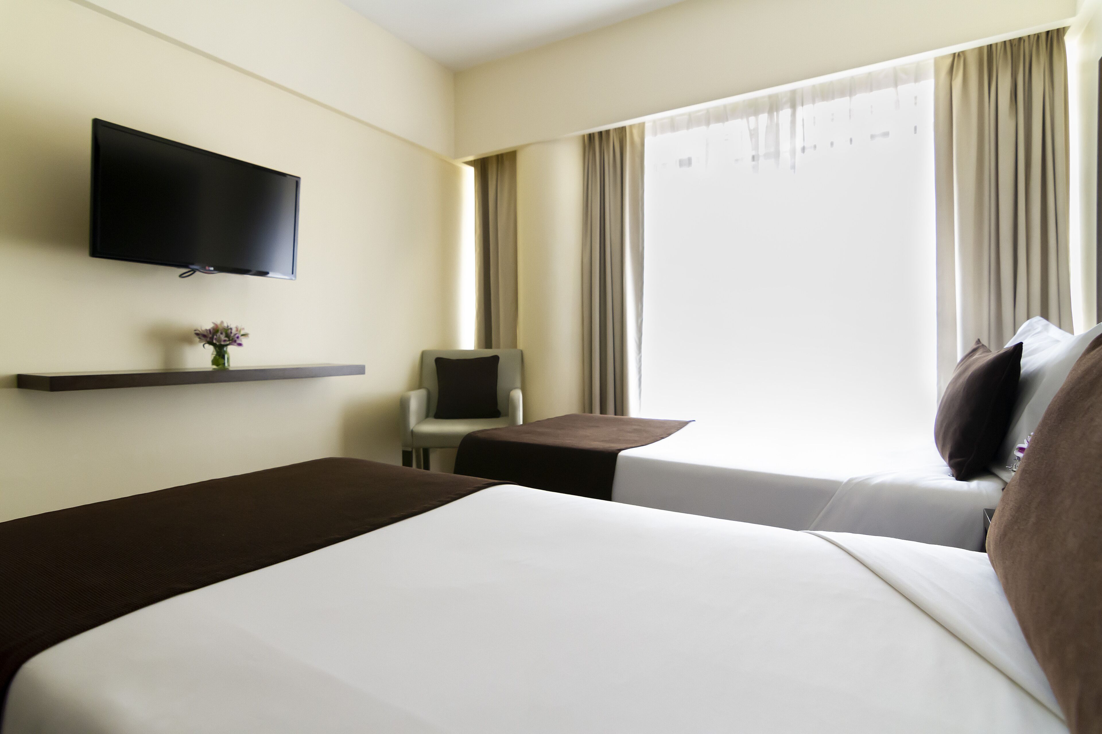 A hotel room with two beds, a TV mounted on the wall, a small table with a chair, and a window with curtains.