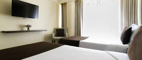 A hotel room with two beds, a TV mounted on the wall, a small table with a chair, and a window with curtains.