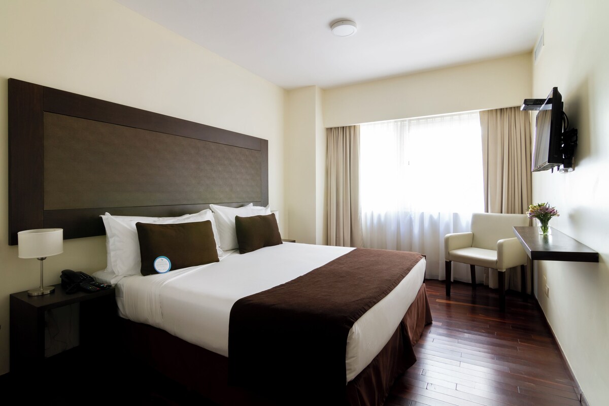 Een hotelkamer met een groot bed, nachtkastje met een lamp, een stoel, bureau met een flatscreen televisie en een raam met gordijnen.