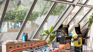 A foosball table in a room with large windows, a small table with books, and a video game machine. - ibis Paris Alésia Montparnasse 14ème (Paris)