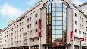 A modern hotel building with a prominent glass corner, multiple balconies, and red signage indicating it is an ibis hotel. - ibis Paris Alésia Montparnasse 14ème (Paris)