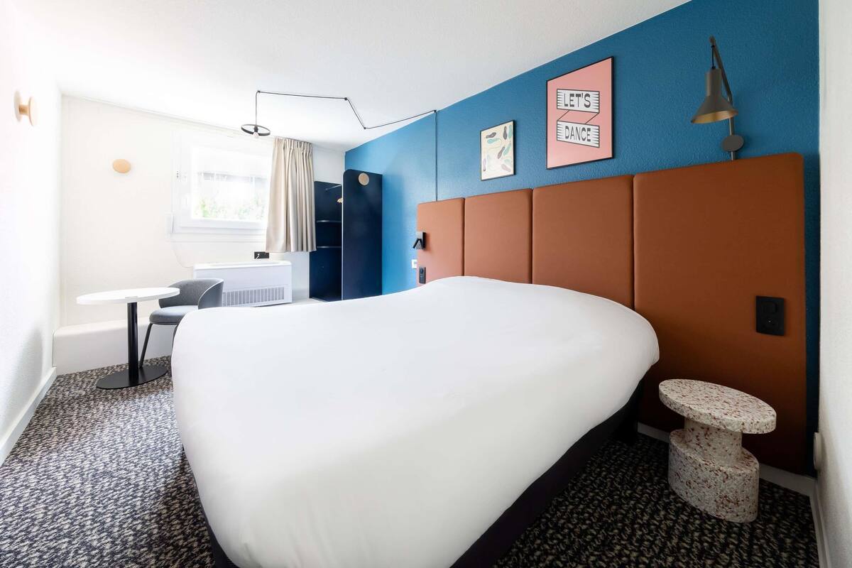 Una habitación de hotel con una cama grande, un escritorio, una silla, una mesita, una lámpara y un cuadro en la pared.