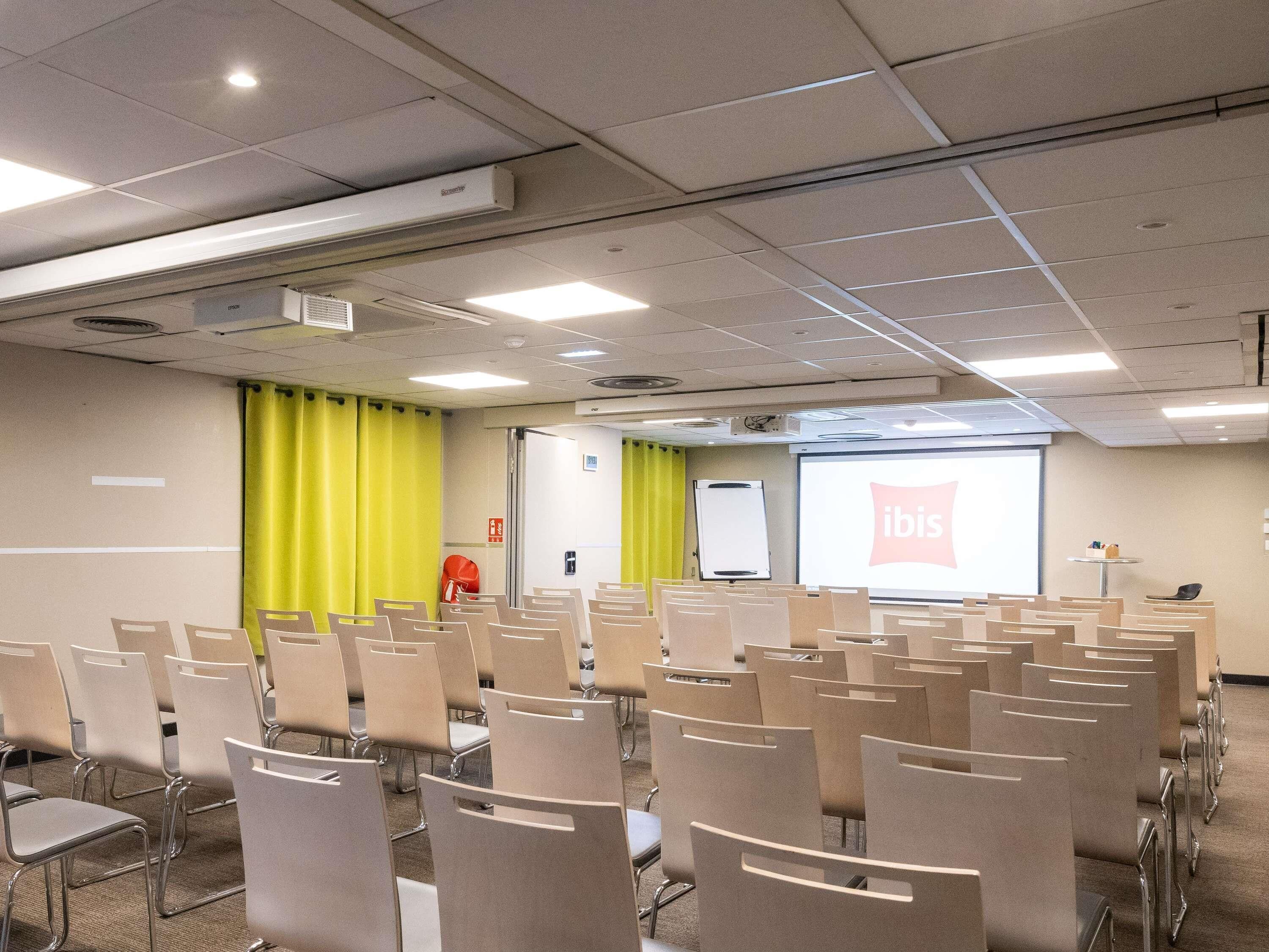 A conference room with rows of chairs, a projector screen displaying 'ibis,' a whiteboard, and a fire extinguisher.