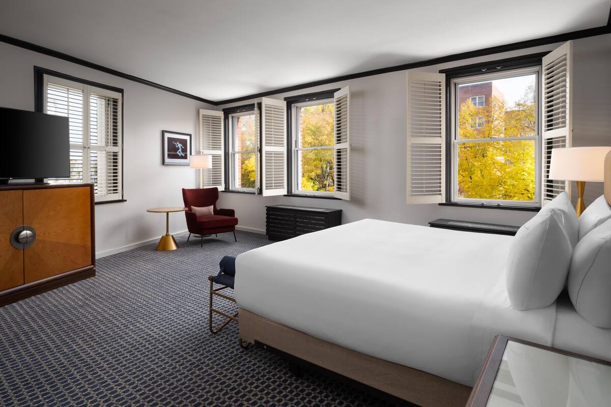 A hotel room with a large bed, a TV, a red chair, and multiple windows with shutters.