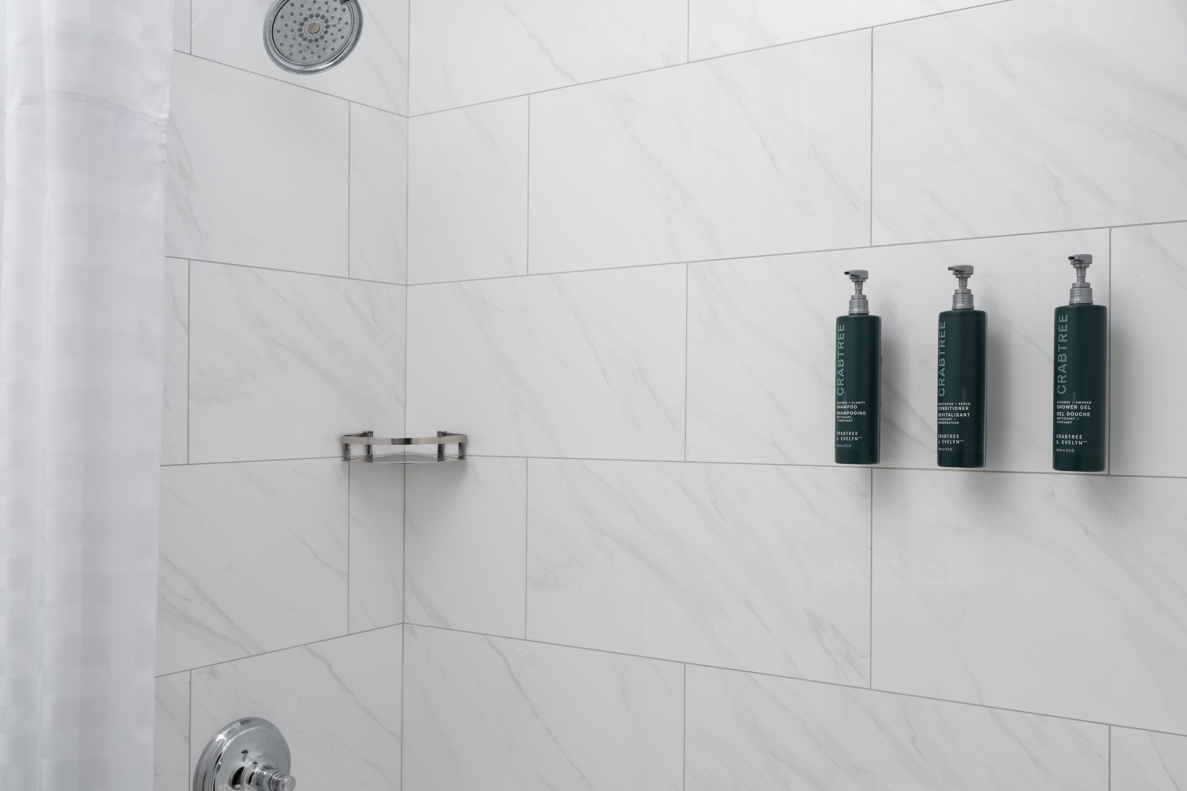 Ein Badezimmer mit weißen Marmorfliesen, einer Duschbrause und drei schwarzen Flaschen mit dem Schriftzug 'Crabtree' an der Wand.