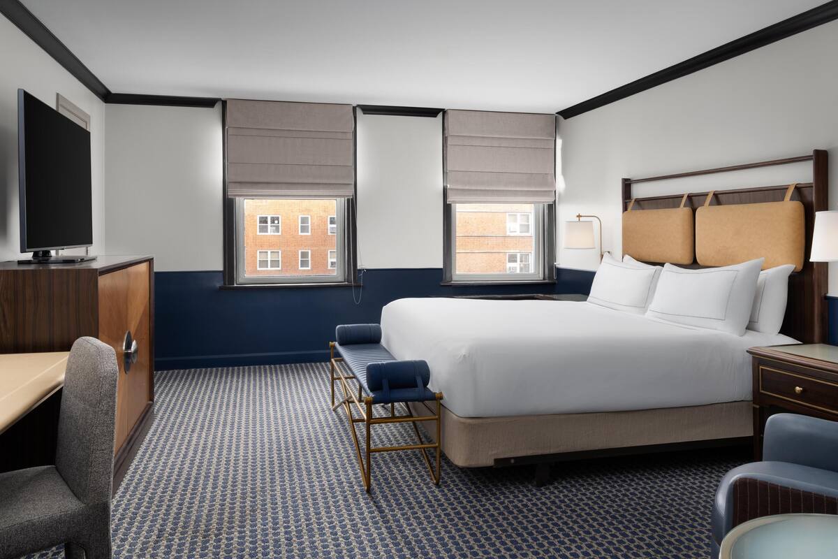 A hotel room with a large bed, a desk, a chair, and two windows with blinds.