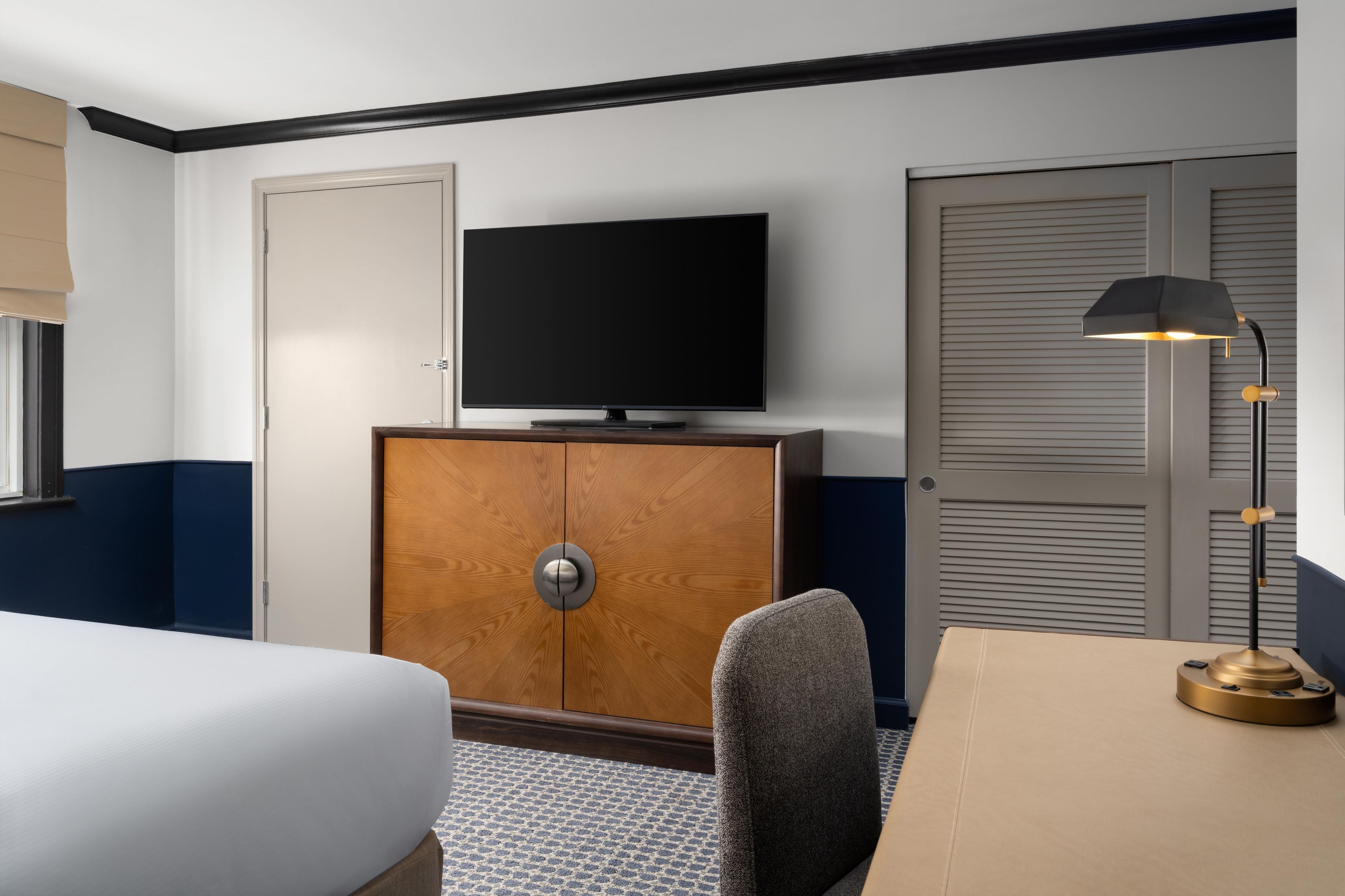 Una habitación de hotel con cama, televisión sobre un mueble de madera, escritorio con lámpara y armario.
