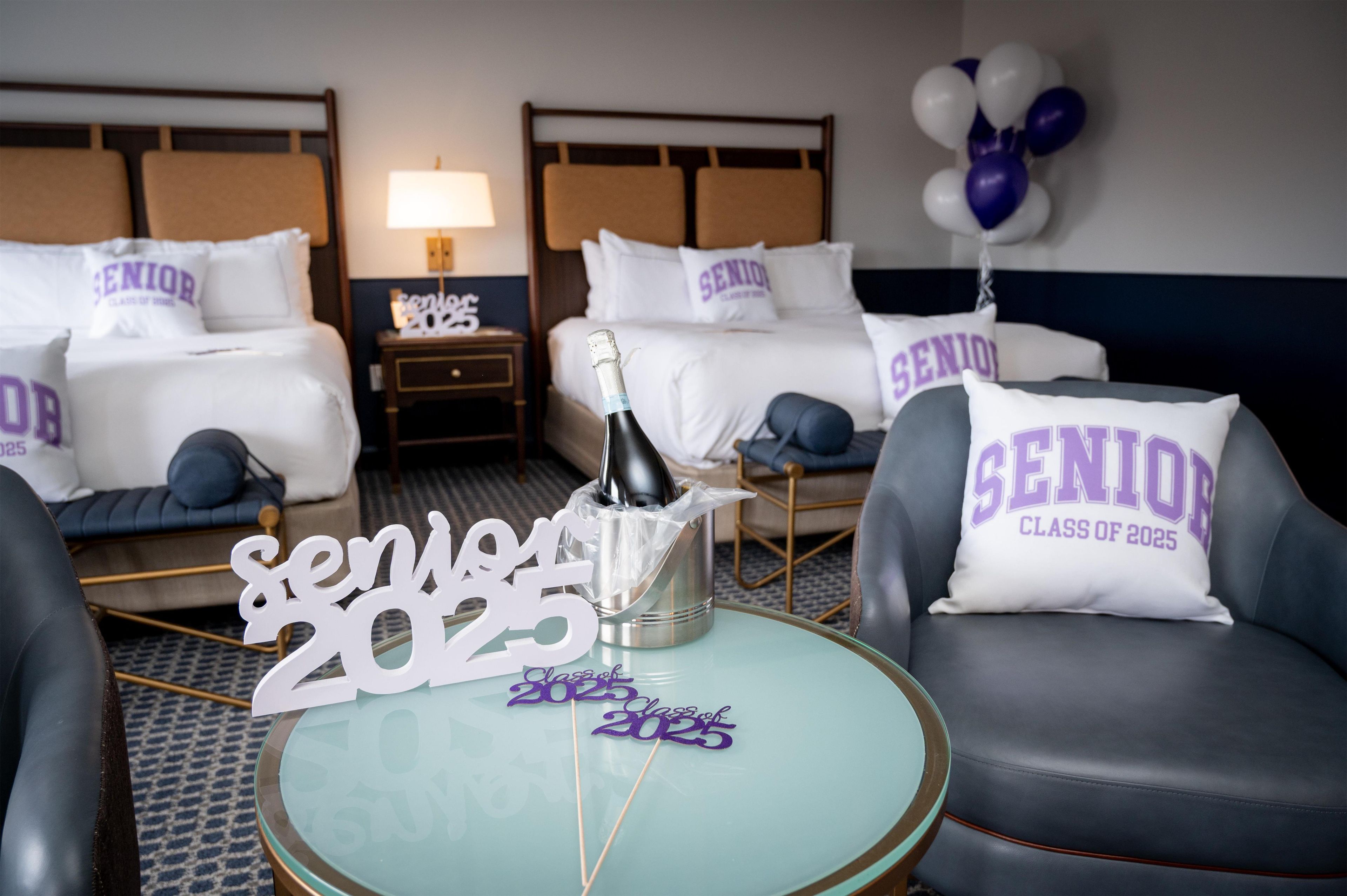 A hotel room with two beds, a nightstand, a round table with a bottle of champagne, and a sign that reads 'SENIOR CLASS OF 2025'.