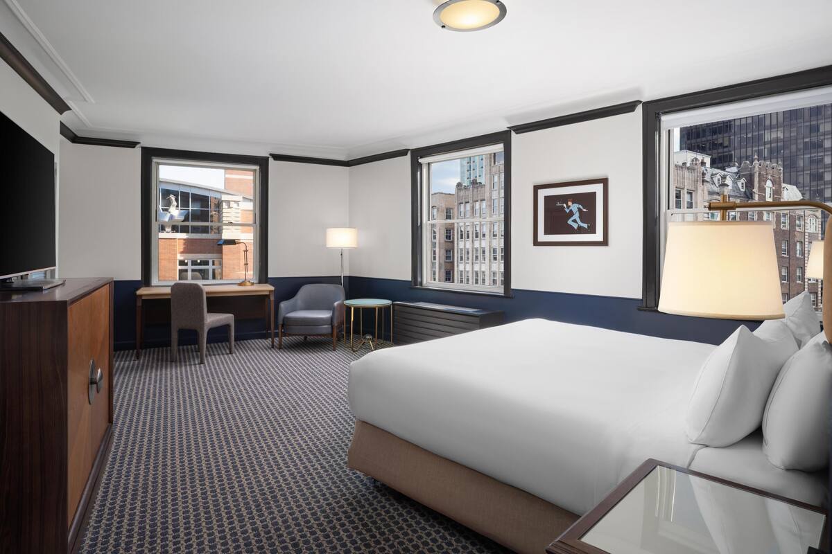 A hotel room with a large bed, a desk with a chair, a small table, and a TV.