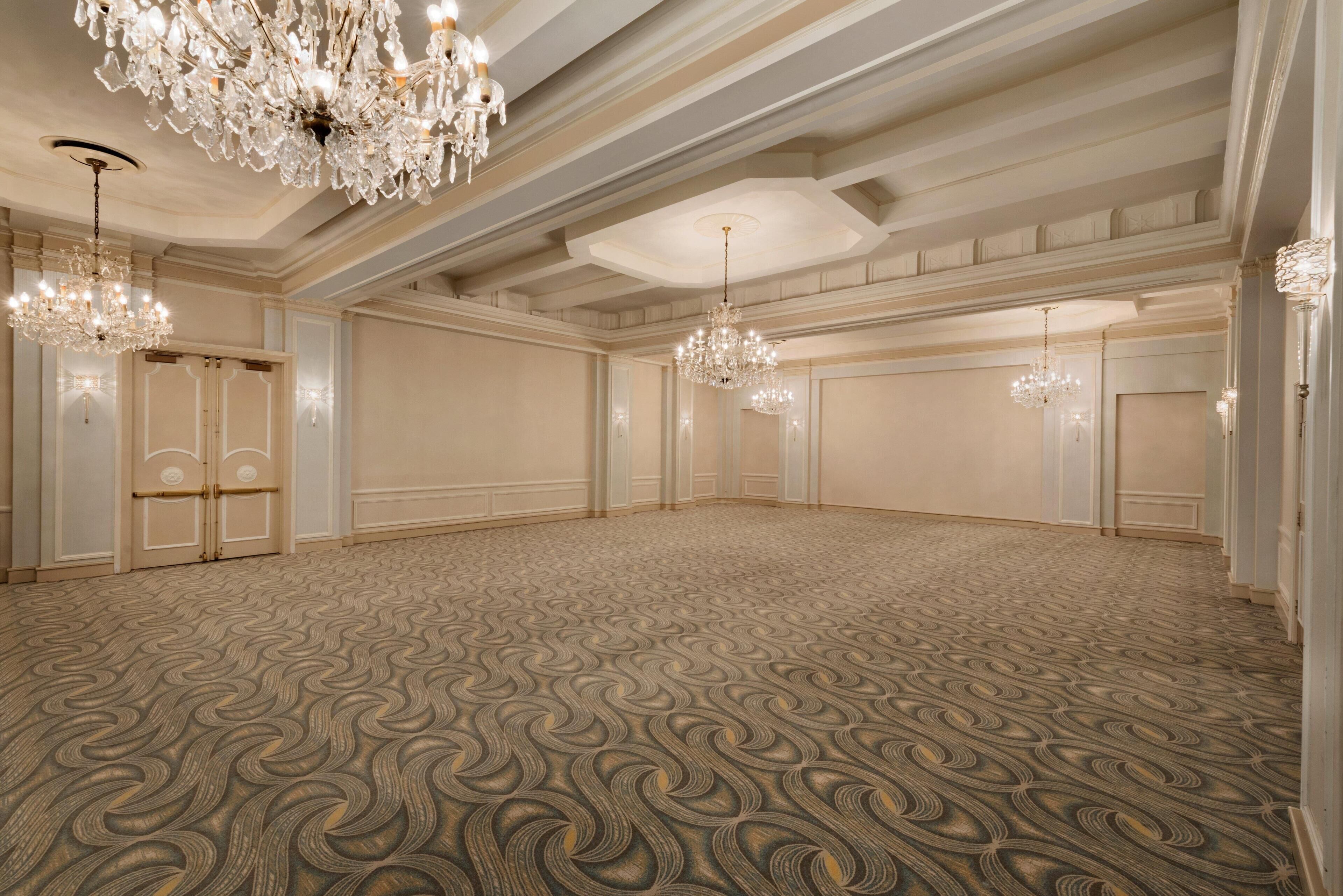 A spacious room with a patterned carpet, ornate chandeliers, and a classic architectural design.