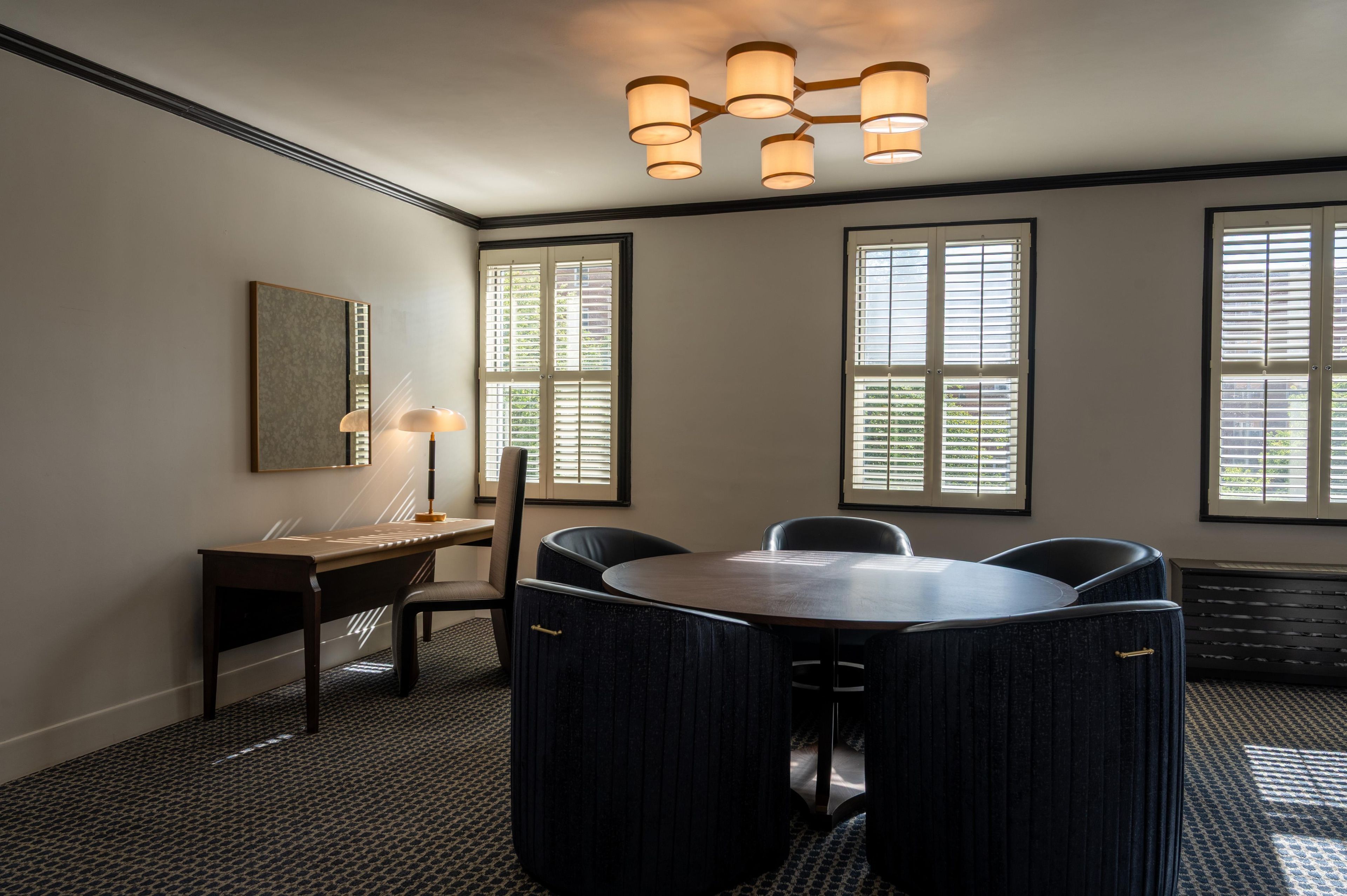 A conference room with a round table, four chairs, a desk with a lamp, and a large window with blinds.