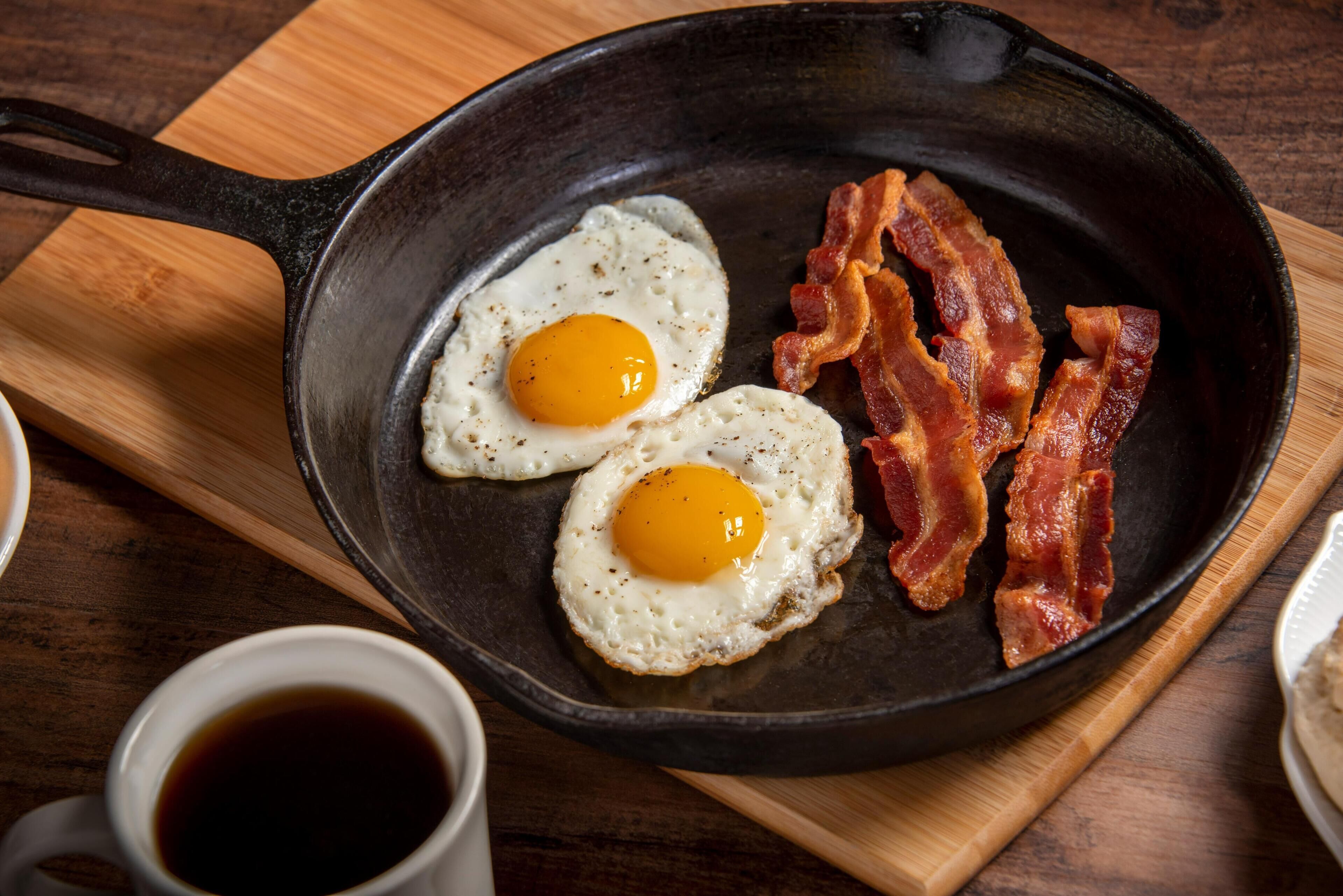 Una sartén con dos huevos estrellados y tiras de tocino sobre una tabla de madera.