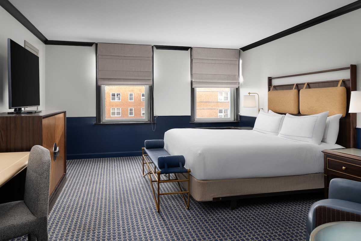 Habitación de hotel con una cama grande, un escritorio, una silla y dos ventanas con persianas.