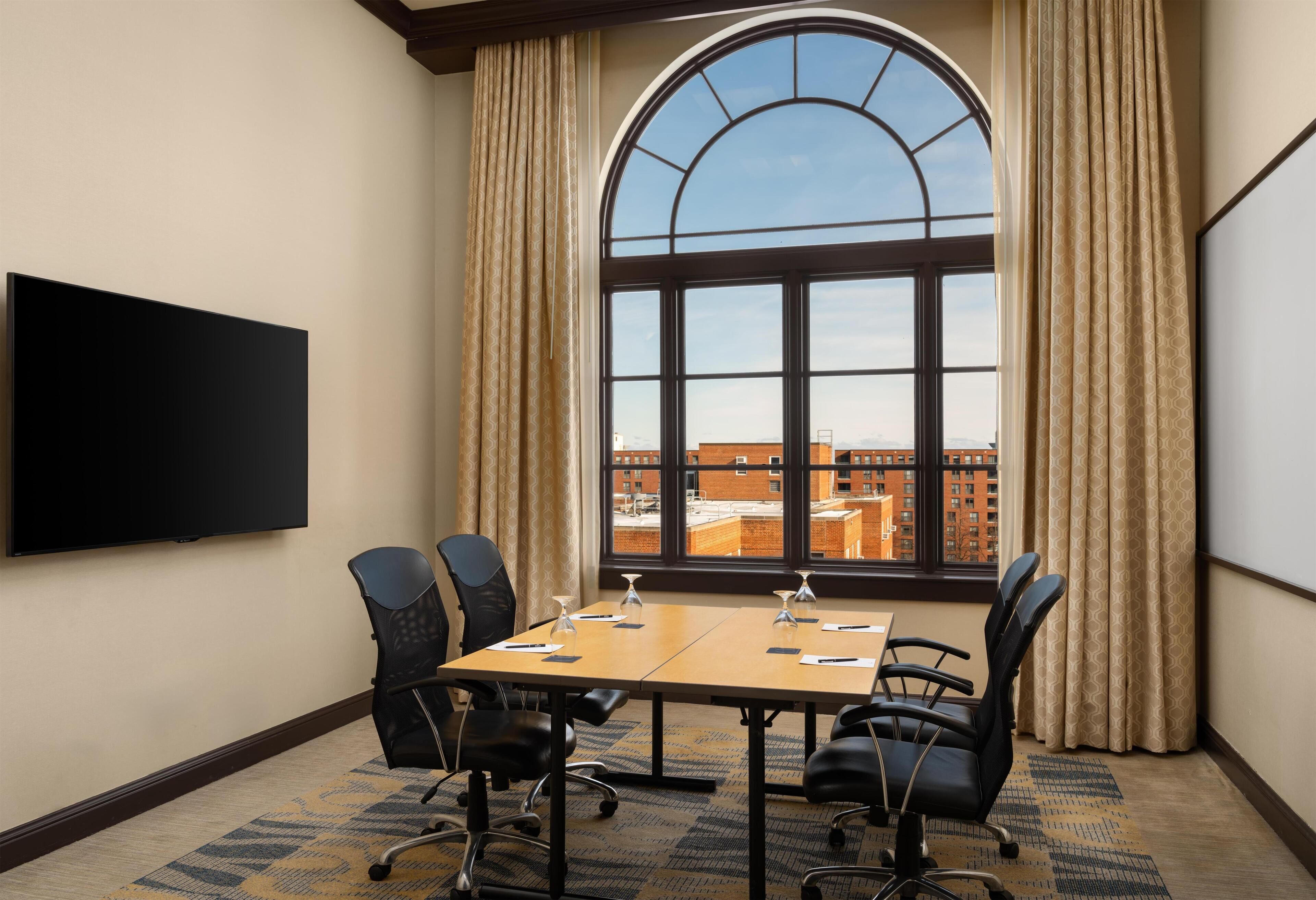A conference room with a large table, four chairs, a flat-screen TV, and a large arched window with curtains.