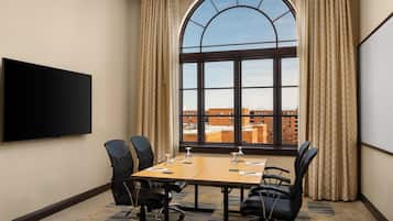 A conference room with a large table, four chairs, a flat-screen TV, and a large arched window with curtains.