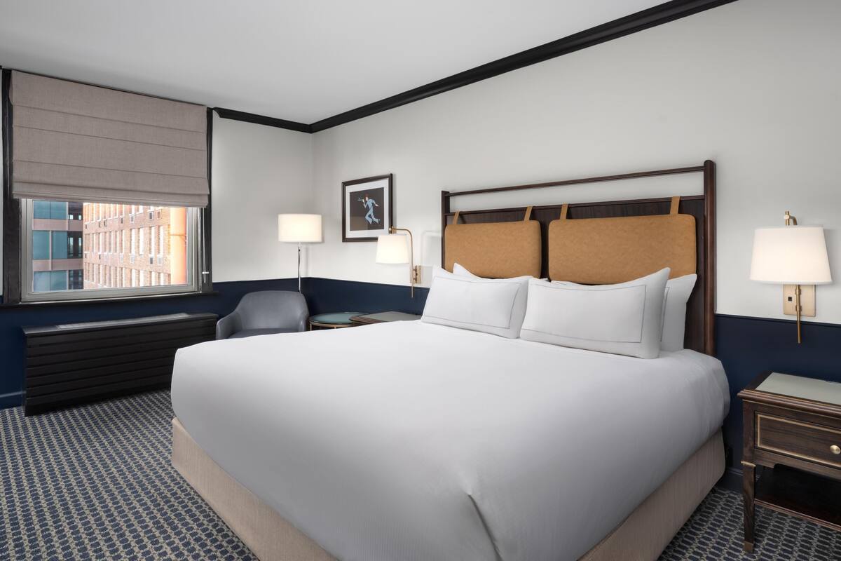 A hotel room with a large bed, a desk, a chair, and a window with a view of buildings.
