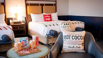 A hotel room with two beds, a round coffee table with gingerbread cookies, and a chair with a pillow labeled 'HOT COCOA'.