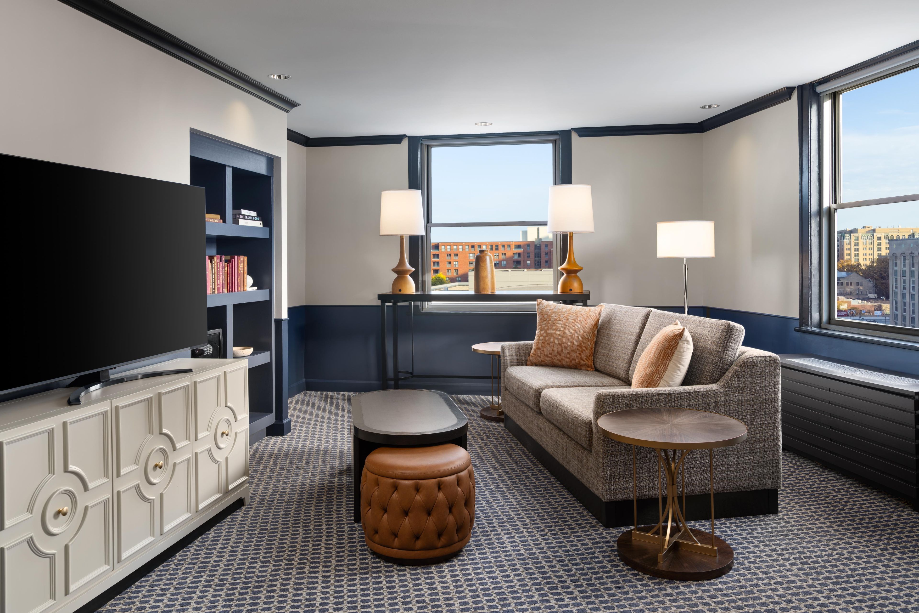 A hotel room with a large flat-screen TV, a sofa, a coffee table, a side table, and a bookshelf.