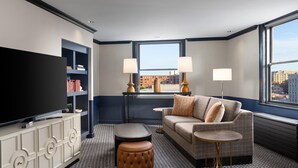 Une chambre d’hôtel moderne dotée d’un grand téléviseur à écran plat, d’un canapé gris agrémenté d’un plaid à motifs, d’une table basse ronde et d’une bibliothèque.