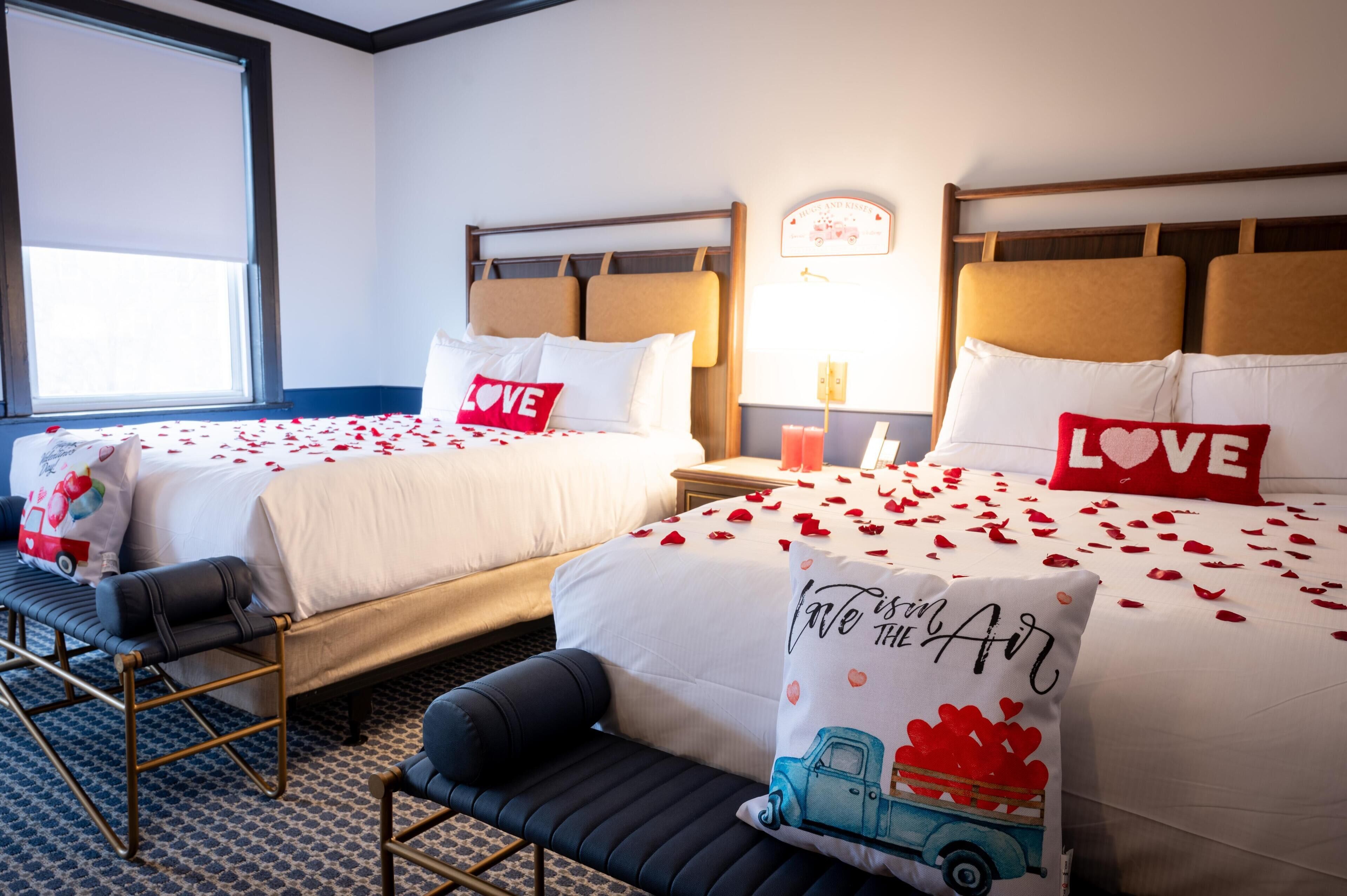 Une chambre d’hôtel avec deux lits, chacun orné d’un coussin sur lequel est brodé le mot « AMOUR ». On y trouve également un banc et une petite table avec une lampe.