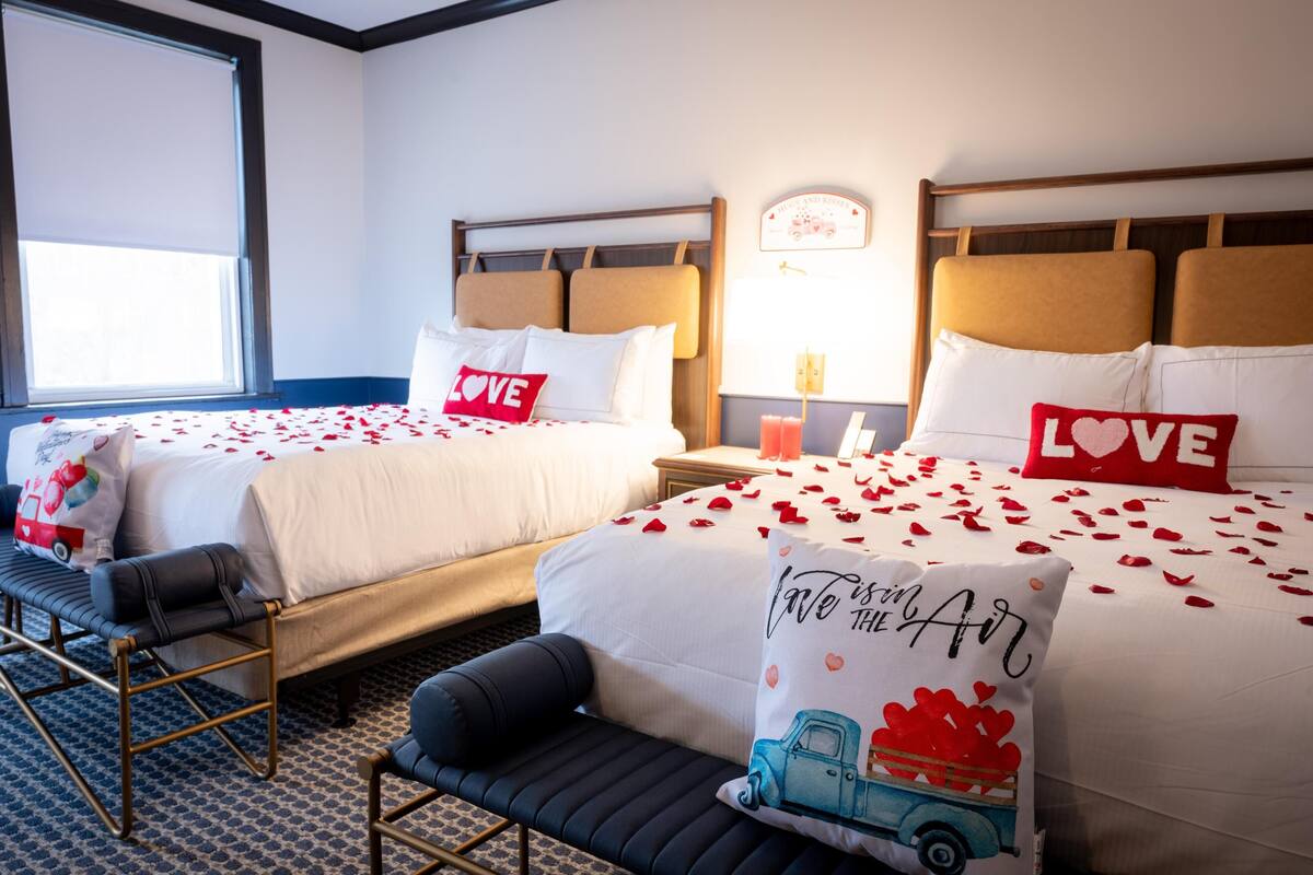 A hotel room with two beds, each with a pillow that says "LOVE". There is a bench and a small table with a lamp.