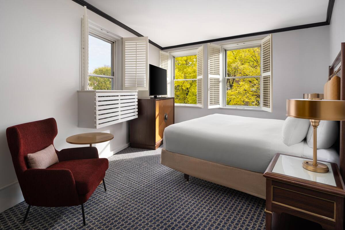 A hotel room with a bed, a red armchair, a TV, and a window with shutters.