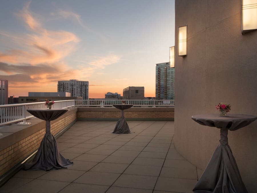 A rooftop terrace with three tables, each with a vase of flowers, set against a backdrop of a city skyline at sunset.