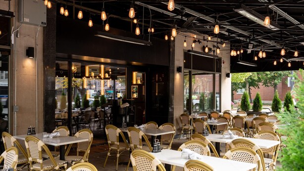 Un espace extérieur aménagé pour les repas, avec des chaises et des tables en osier, un éclairage suspendu et une vue sur la rue.