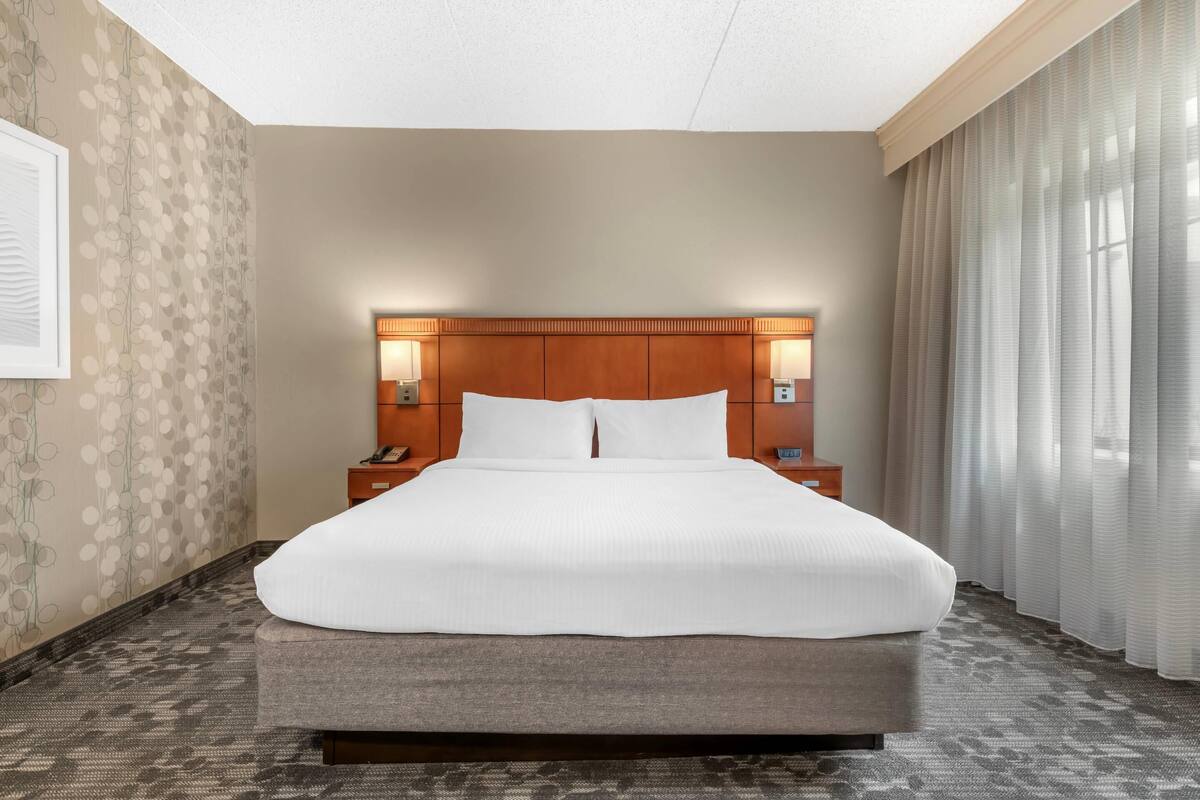 Una habitación de hotel con una cama grande, cabecero de madera, lámparas de noche y alfombra estampada.