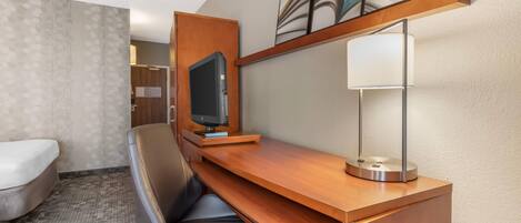 A hotel room with a wooden desk, a television, a lamp, a chair, and a bed.