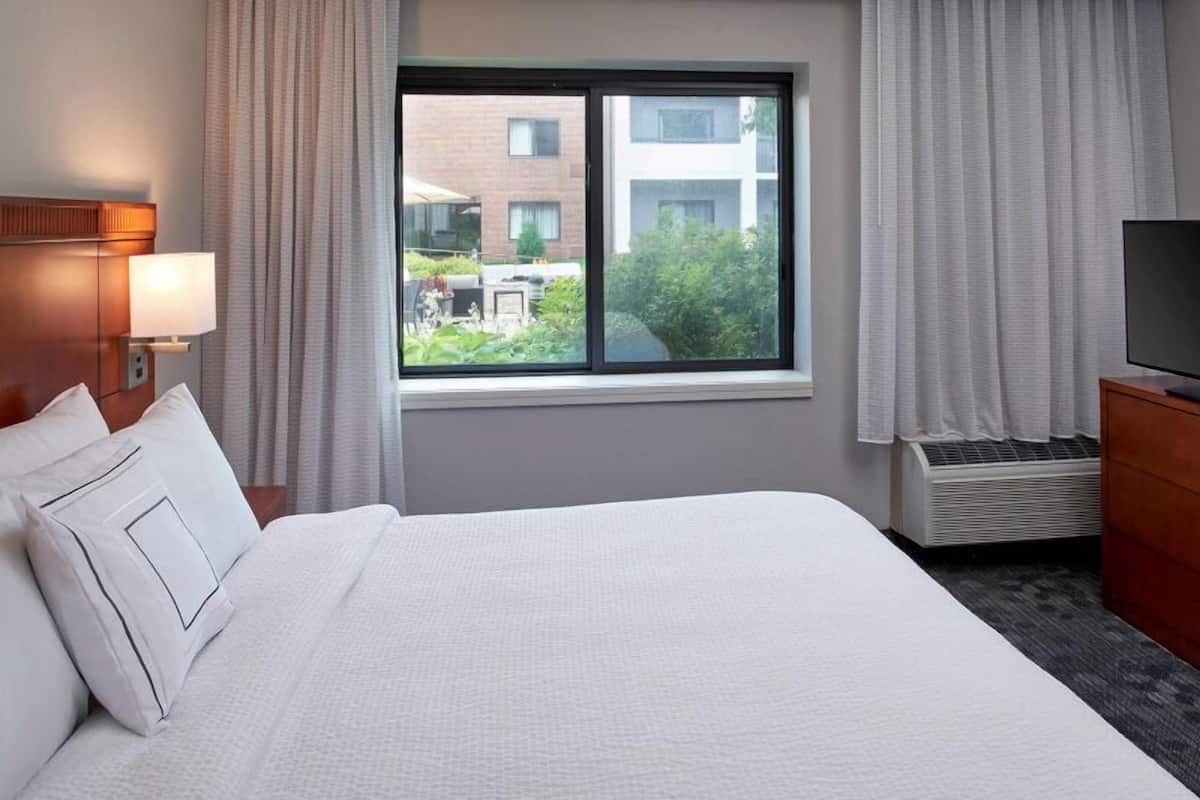 Habitación de hotel con cama, televisor, ventana con cortinas y vistas a un edificio y vegetación.