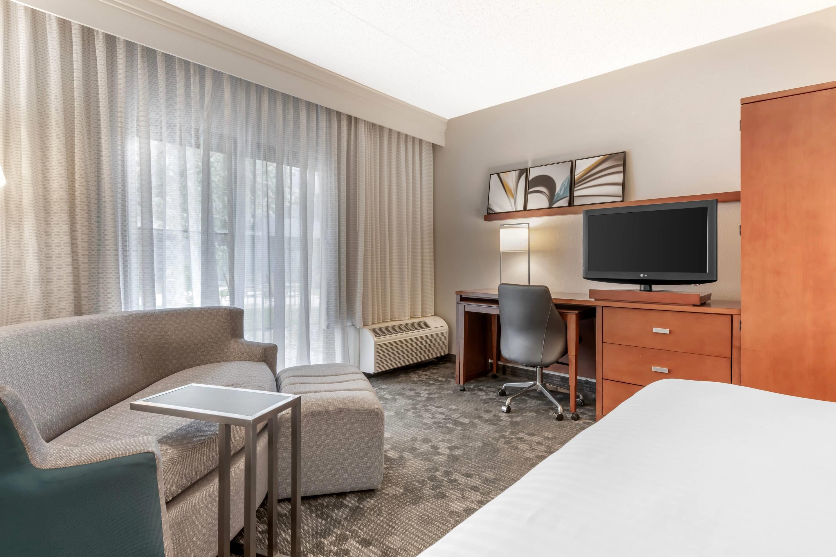 A hotel room with a sofa, a small table, a desk with a chair, a TV, and a wardrobe.