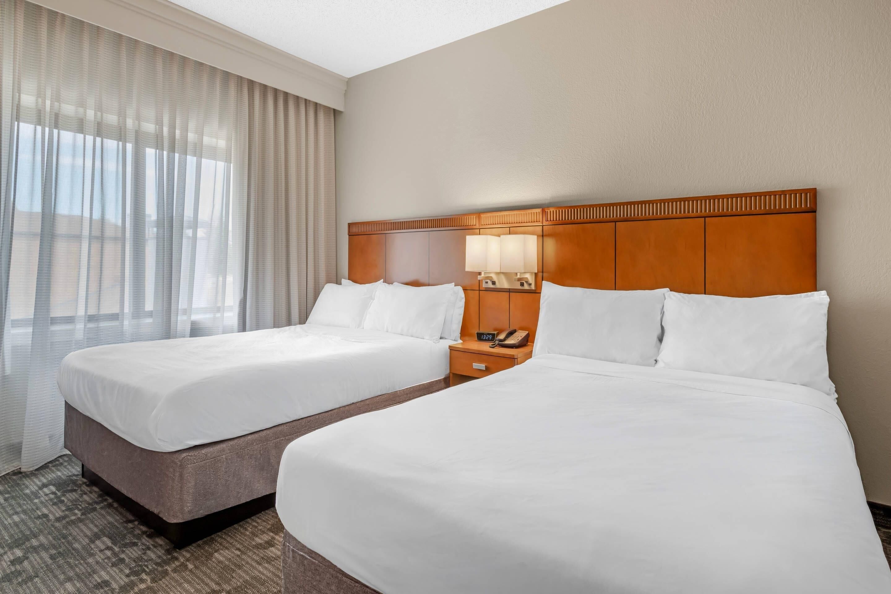A hotel room with two beds, a nightstand with a lamp, and a window with sheer curtains.