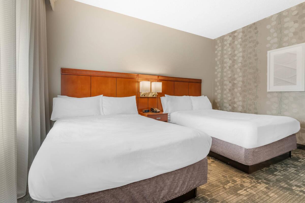 Una habitación de hotel con dos camas, cabecera de madera, mesita de noche con lámpara y pared con diseño.
