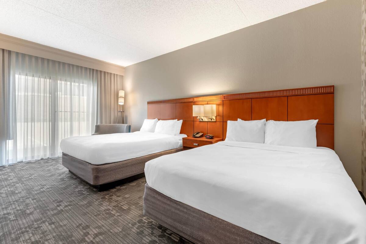 Habitación de hotel con dos camas, un ventanal con cortinas, una mesita de noche con lámpara y un teléfono.