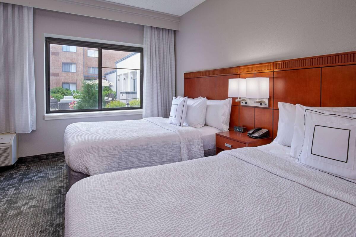 Habitación de hotel con dos camas, una ventana con cortinas, cabecero de madera, mesita de noche con teléfono y vistas a edificios.