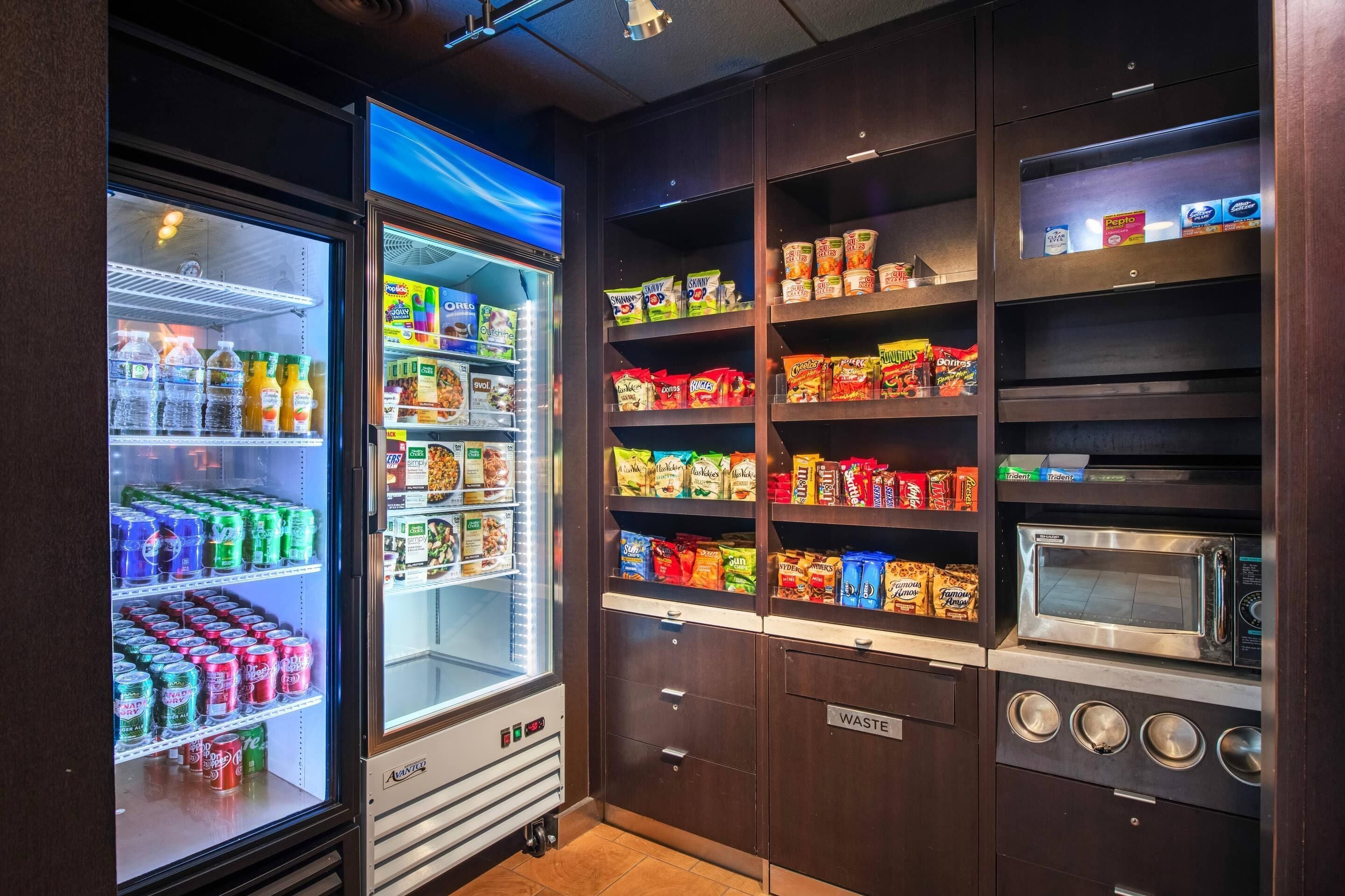 A kitchenette with a refrigerator, microwave, and shelves stocked with snacks and beverages.