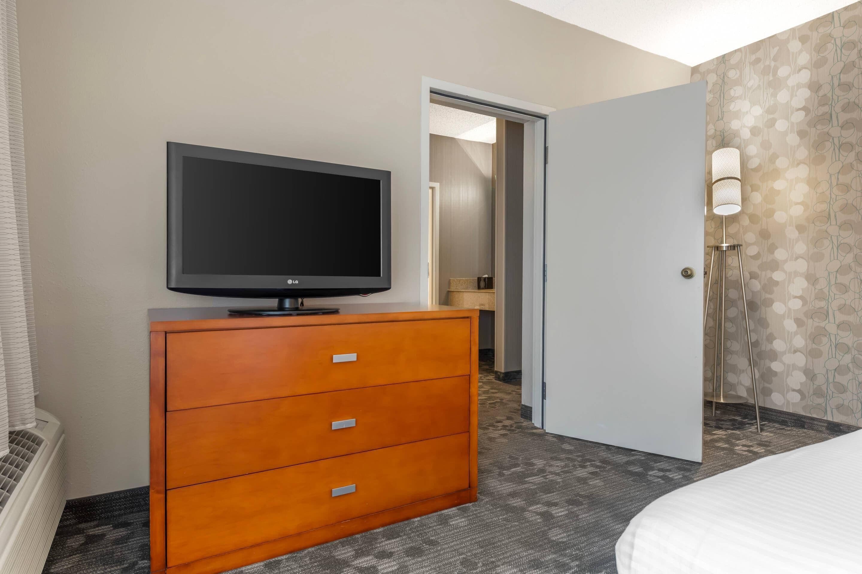 A hotel room with a flat-screen TV on a wooden dresser, a bed, a door leading to a bathroom, and a patterned wall.