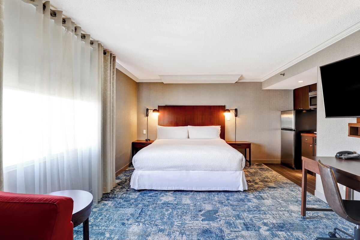 A hotel room with a large bed, a desk, a chair, a television, and a kitchenette.