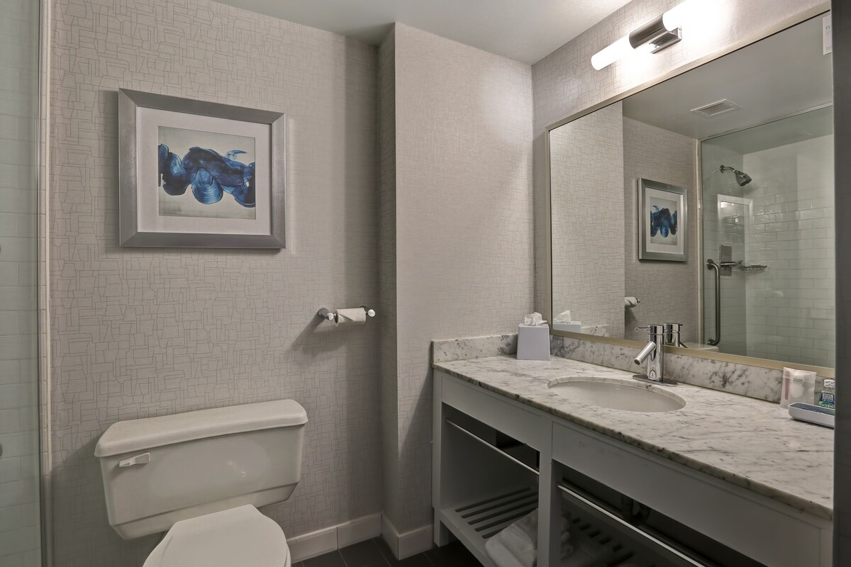 A modern bathroom with a white tiled wall, a marble sink, a toilet, and a framed artwork above the sink.