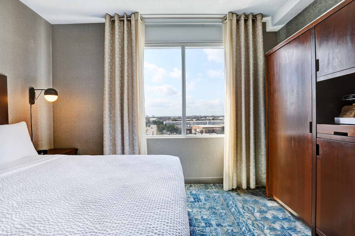 A hotel room with a large bed, a nightstand, a lamp, a wardrobe, and a window with curtains.