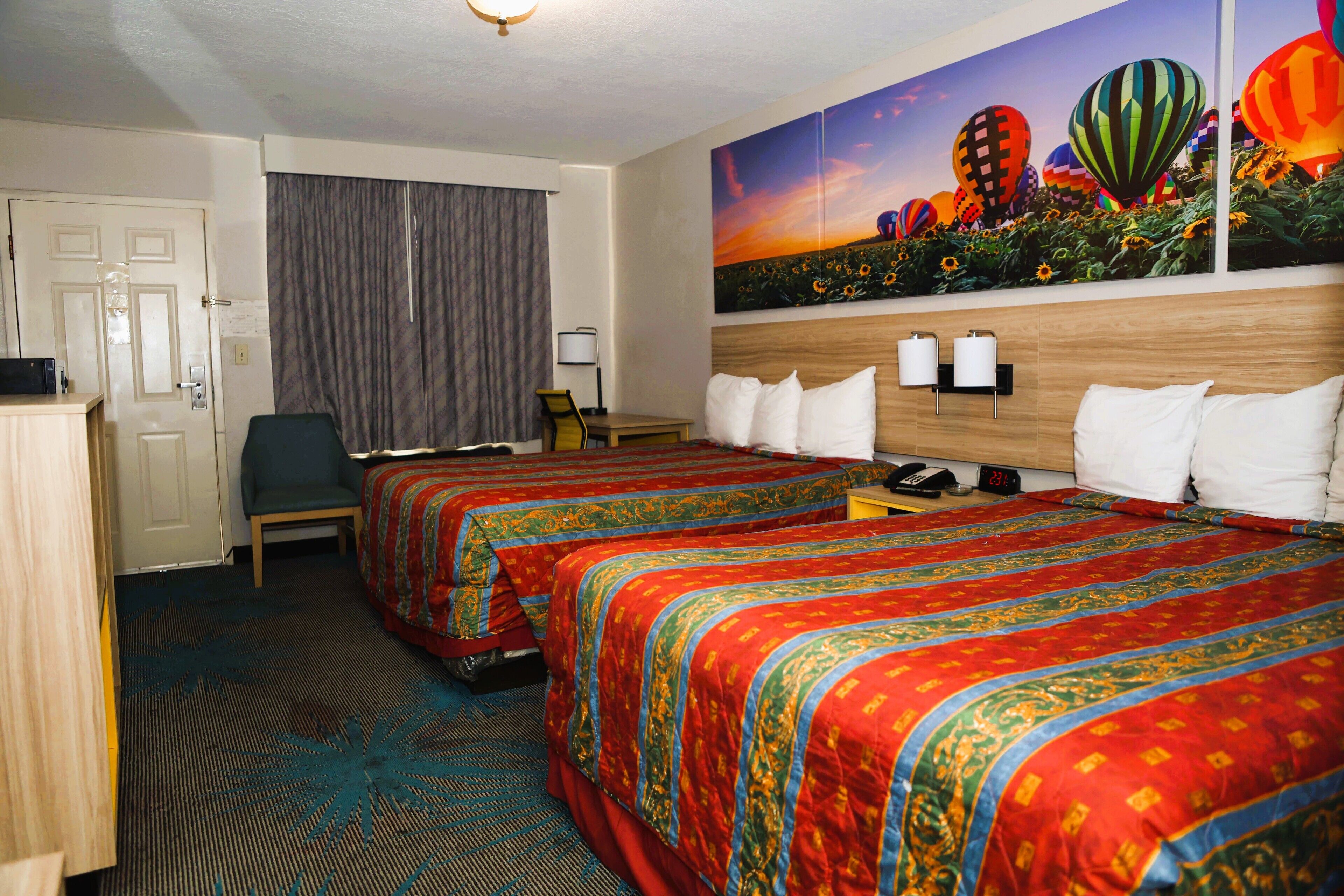 A hotel room with two beds, a desk, a chair, a painting of hot air balloons, and a door.