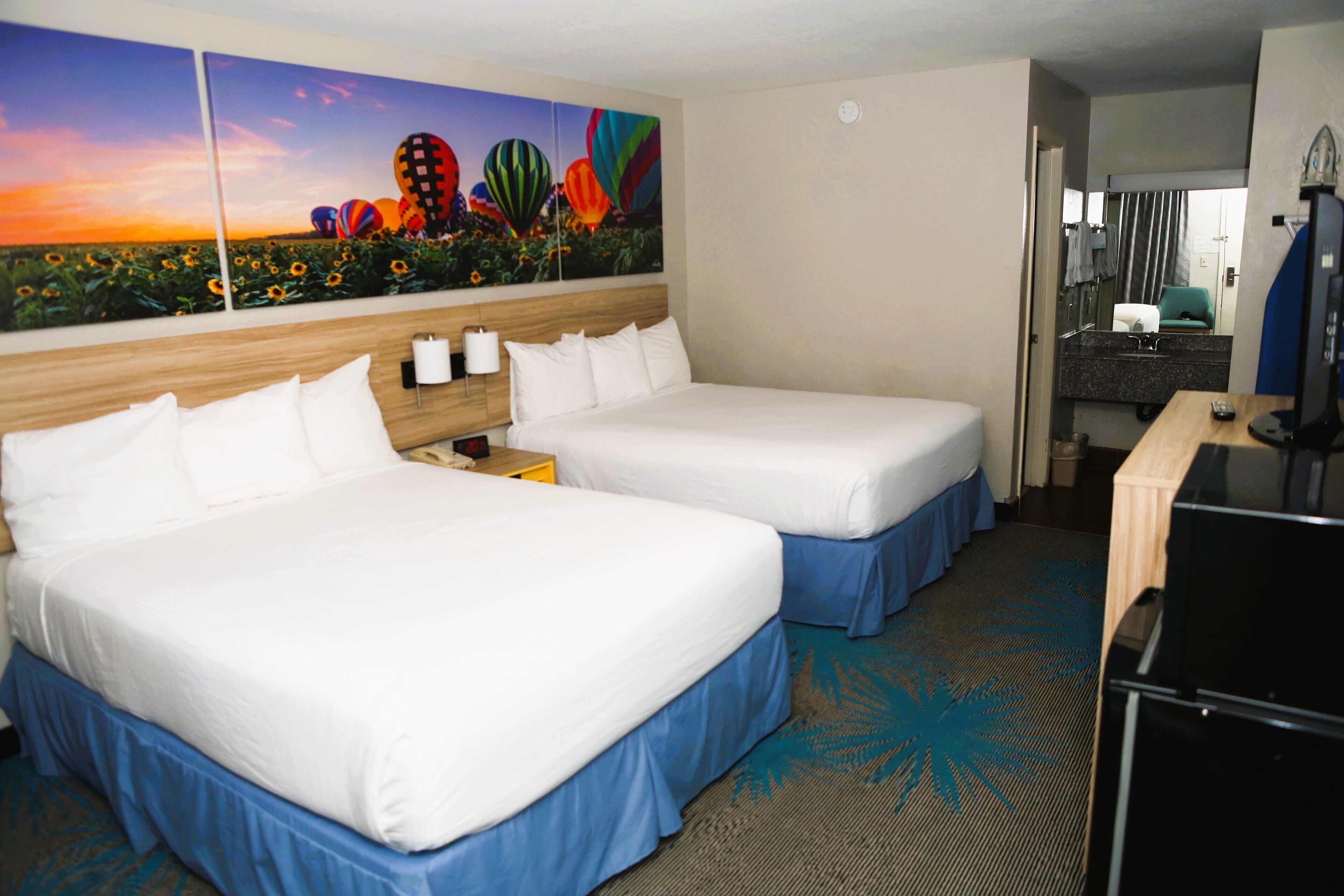 A hotel room with two beds, a painting of hot air balloons, a desk with a computer, and a bathroom visible through an open door.