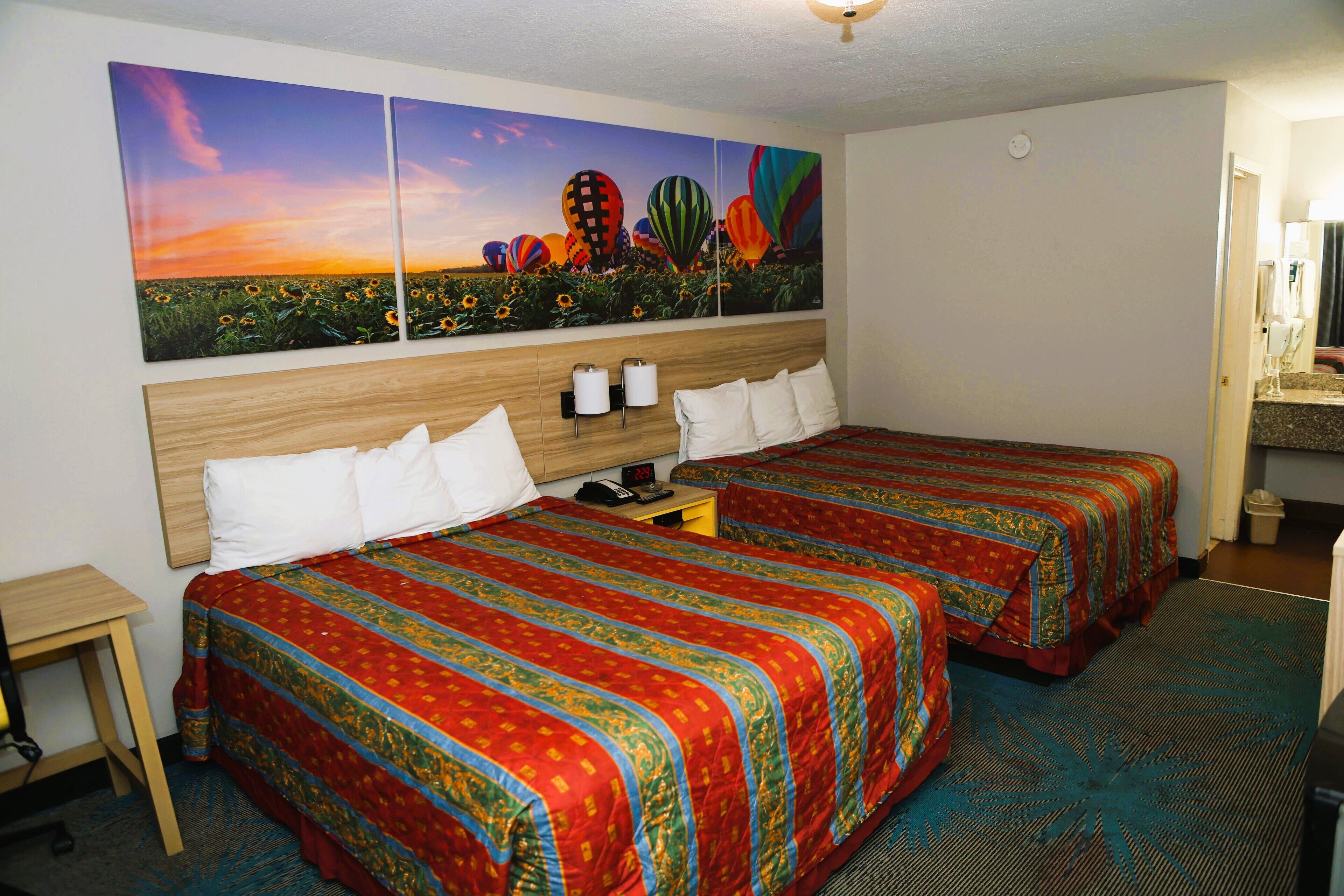 A hotel room with two beds, a large painting of hot air balloons, a wooden headboard, and a small table with a lamp.