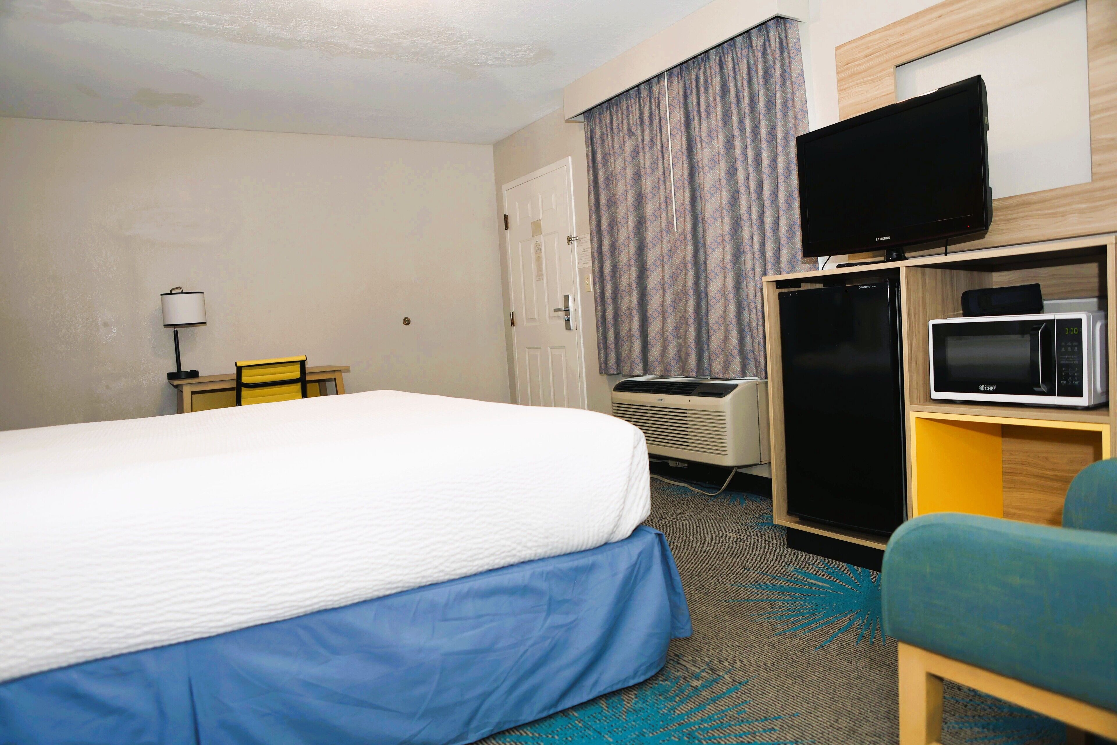 Quarto de hotel com cama, televisão sobre suporte, micro-ondas e pequena escrivaninha com luminária.