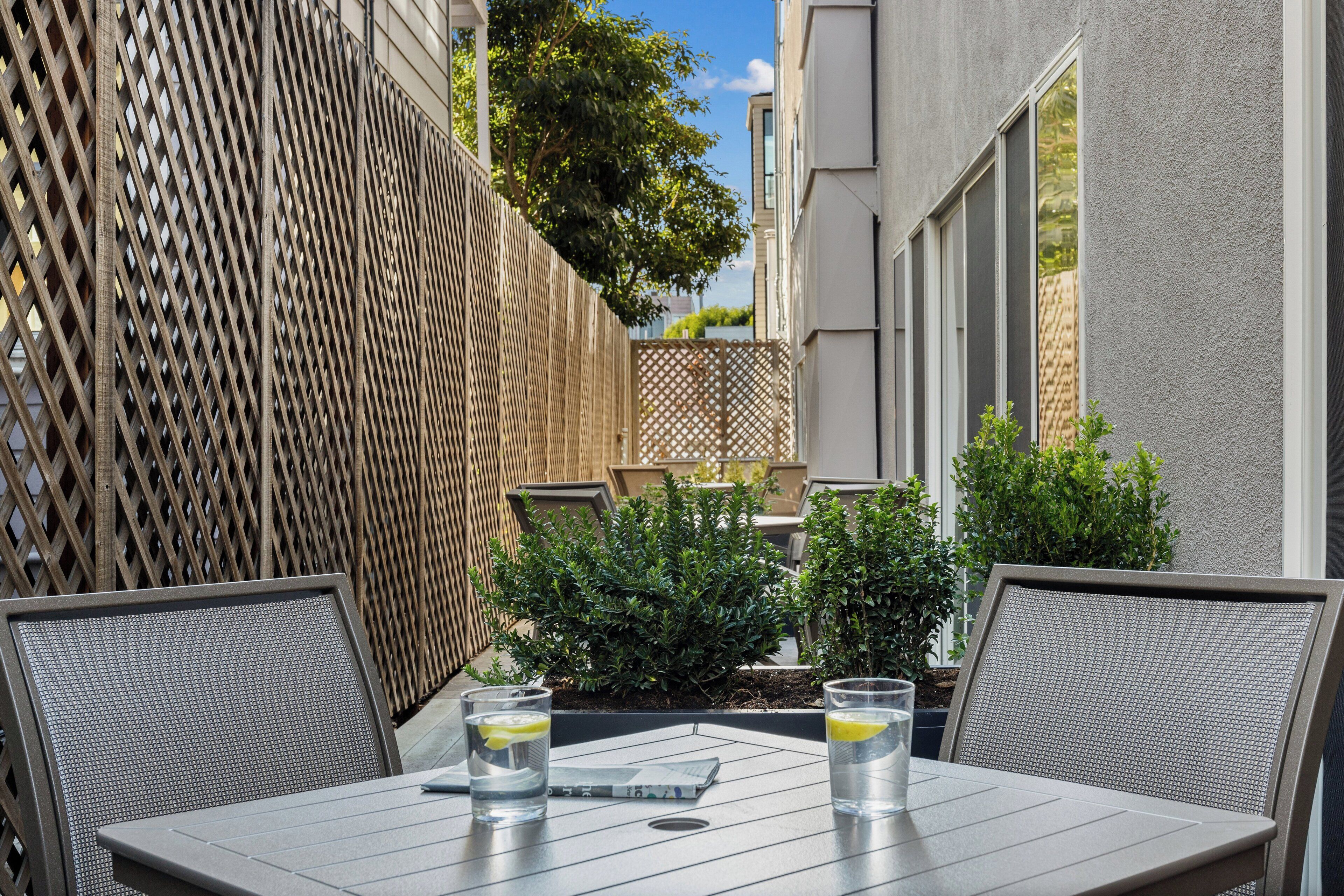Une terrasse avec deux tables, chacune accompagnée d’une chaise, et deux verres d’eau garnis de rondelles de citron. On aperçoit, à l’arrière-plan, des plantes en pot et une clôture en treillis de bois.
