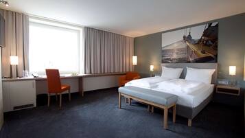 A hotel room with a bed, desk, two chairs, a window with curtains, a nightstand, and a wall-mounted lamp.